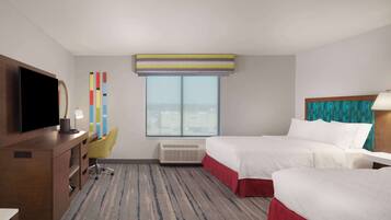 A hotel room with a large bed, a smaller bed, a desk with a flat-screen TV, a chair, a window with a view, and a colorful wall decoration.