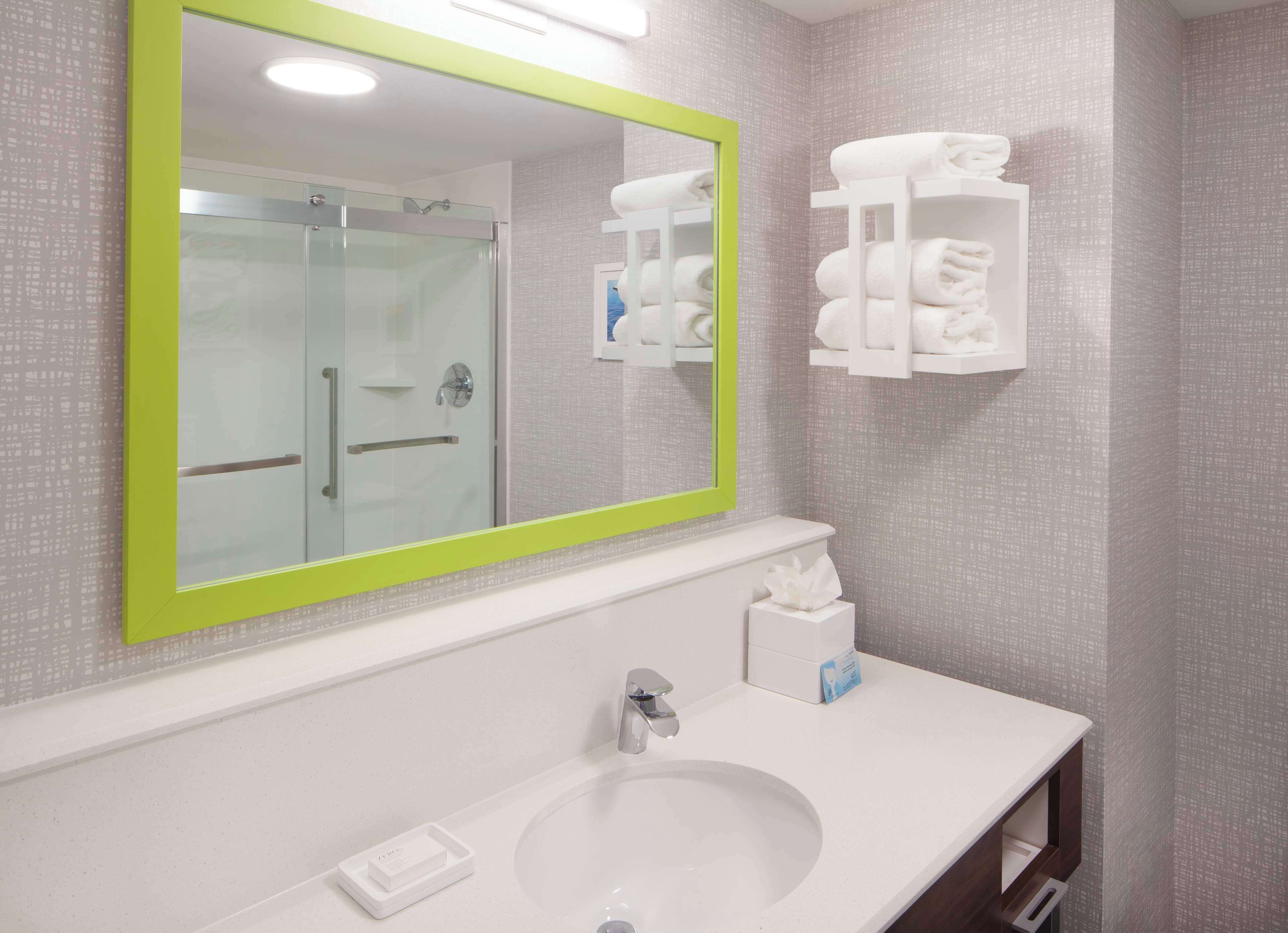 Un baño con un espejo grande, cabina de ducha de vidrio, lavamanos con jabonera y toallero con toallas.