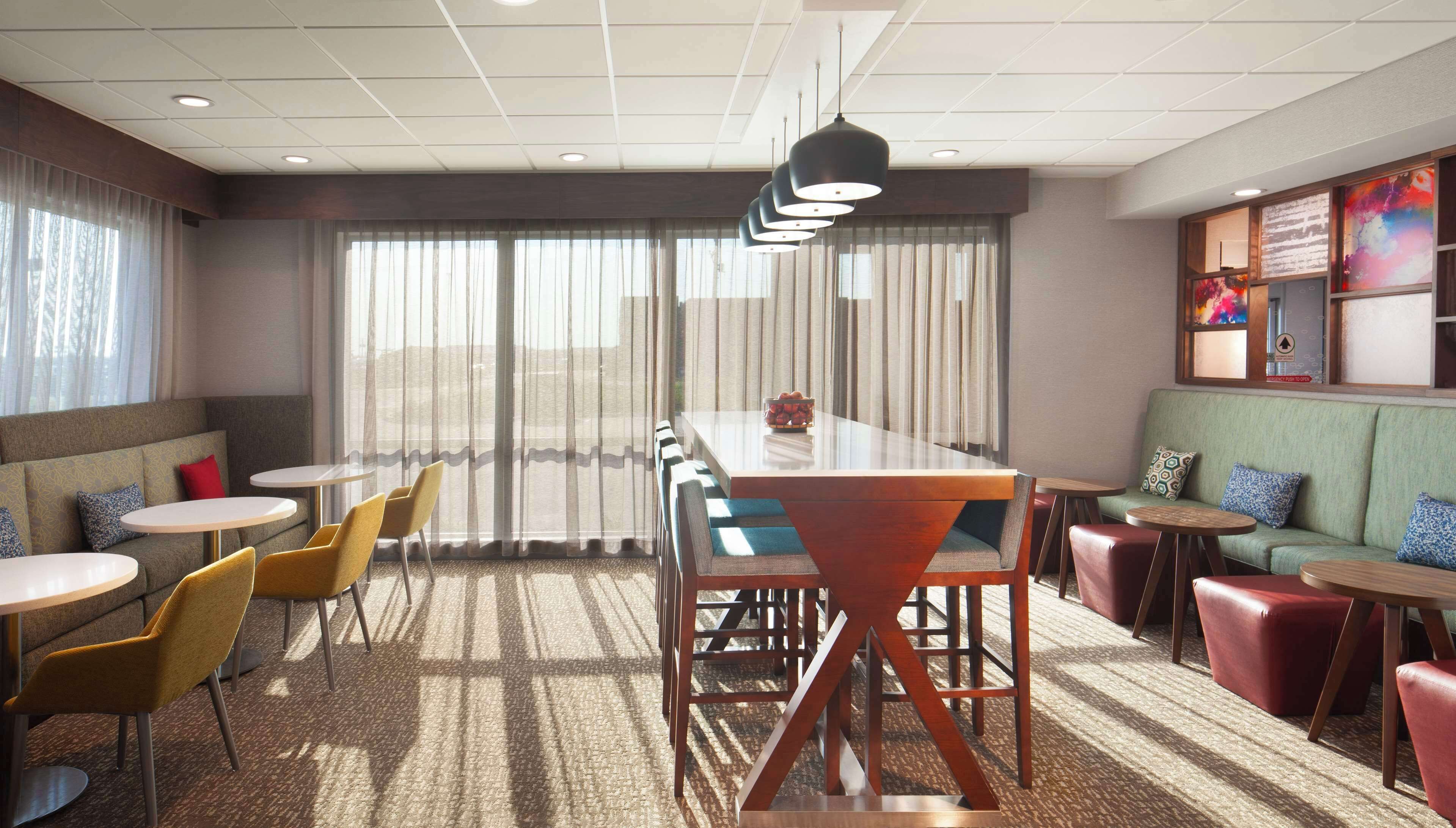 A modern hotel room with a long dining table, multiple seating options including a sofa and chairs, and a large window with sheer curtains.