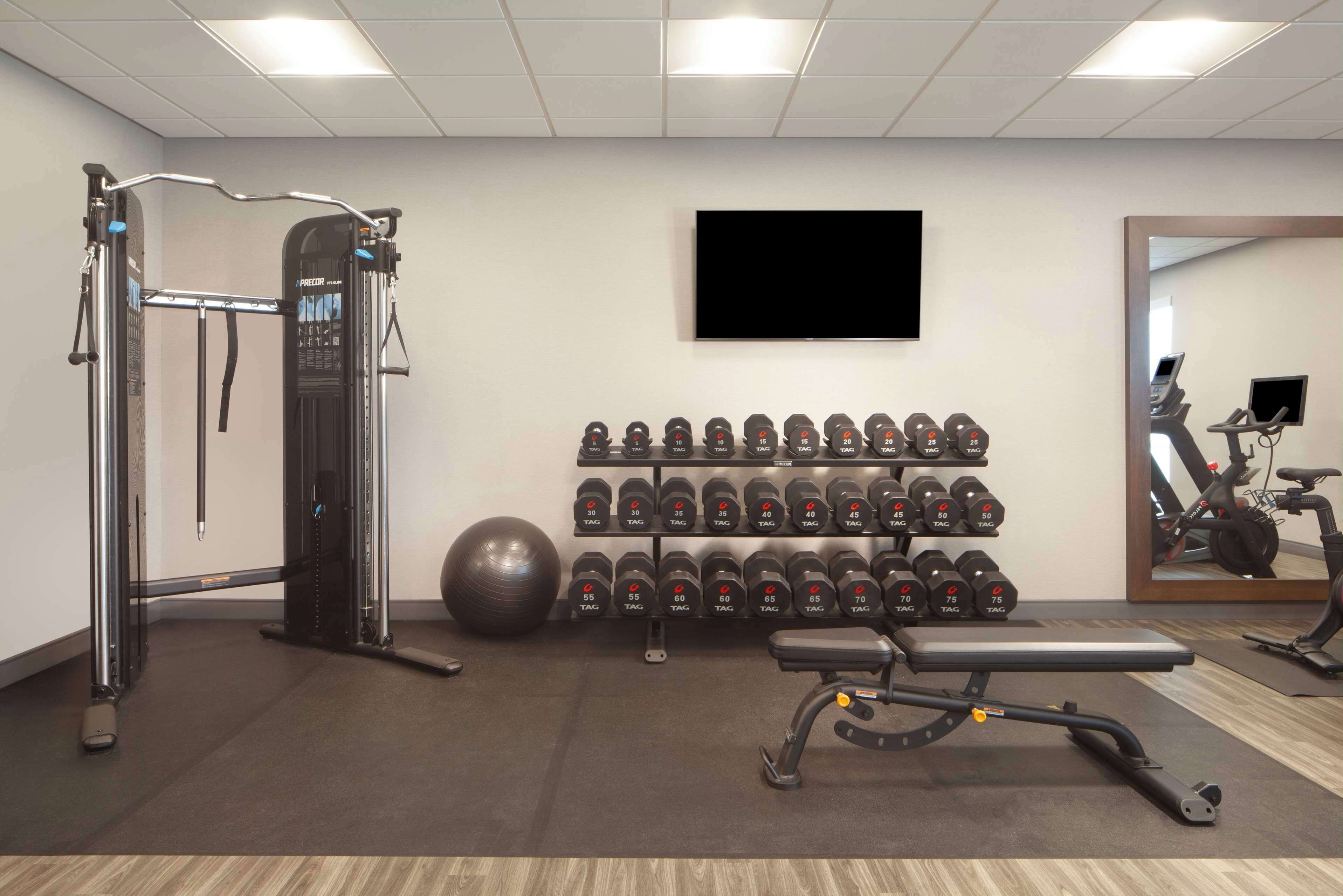 Un gimnasio con equipo de ejercicio, un televisor montado en la pared y un estante con mancuernas.