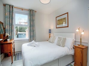 Een opgemaakt bed met witte lakens, een nachtkastje met een lamp en een raam met behang.