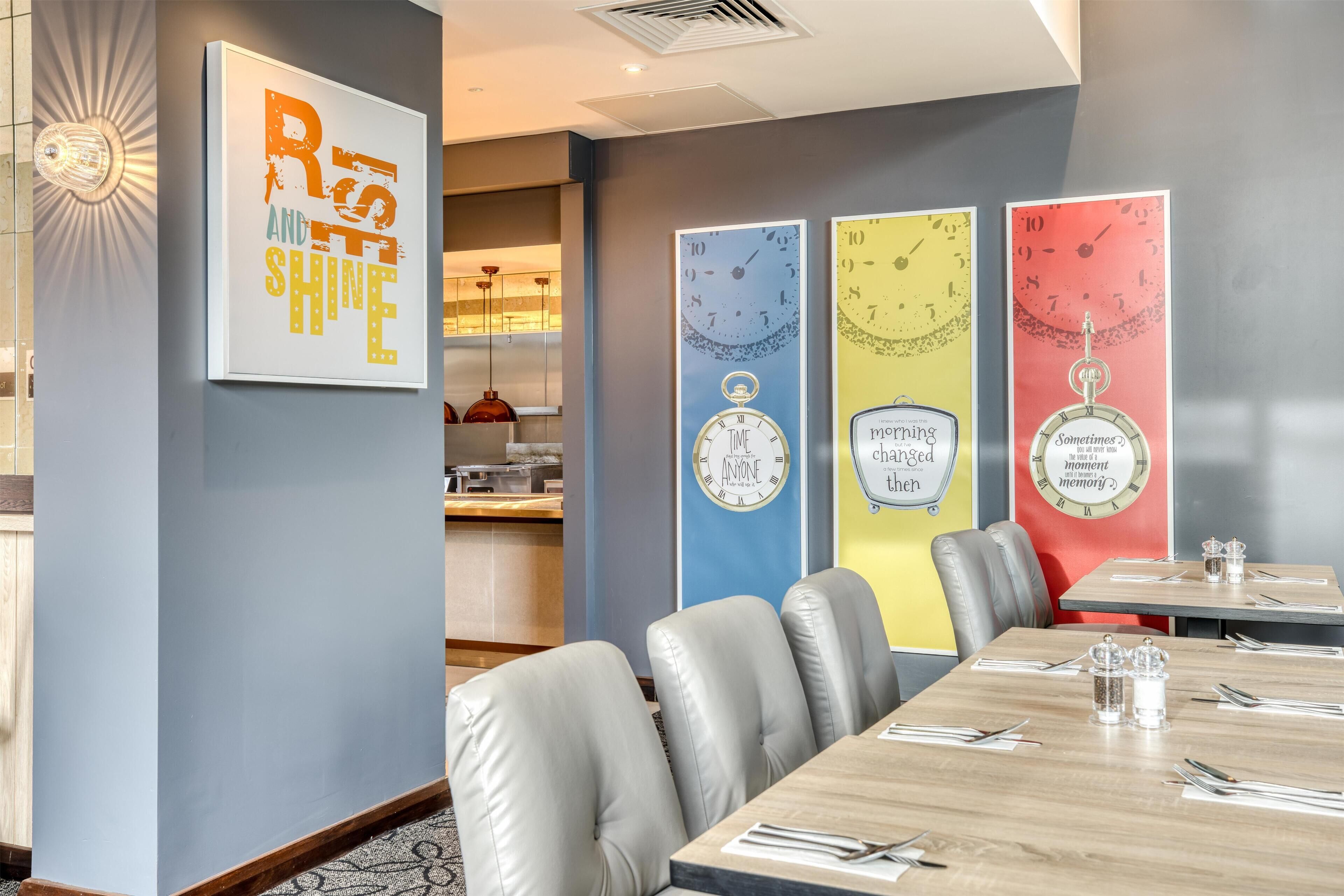 Un comedor con una mesa larga, sillas grises tapizadas y obras de arte que muestran relojes y frases motivacionales.