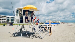 A lifeguard tower with a yellow canopy, situated on a sandy beach with blue umbrellas and lounge chairs in the background. - 2BD/2BA Cocoa Beach Oceanfront + POOL & ALL of Downtown! (Cocoa Beach)