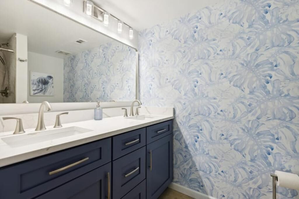 A bathroom with a double sink vanity, dark blue cabinets, and a floral wallpaper pattern.