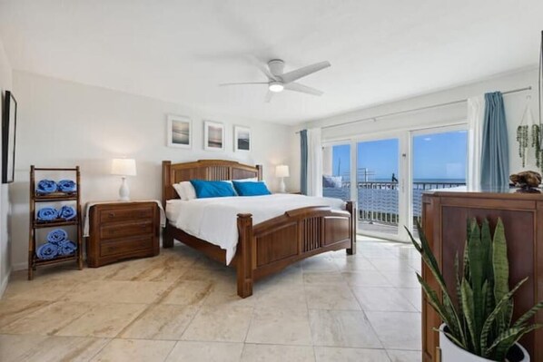 Un dormitorio con cama de madera, una mesita de noche, un aparador y un amplio ventanal con vista al océano.