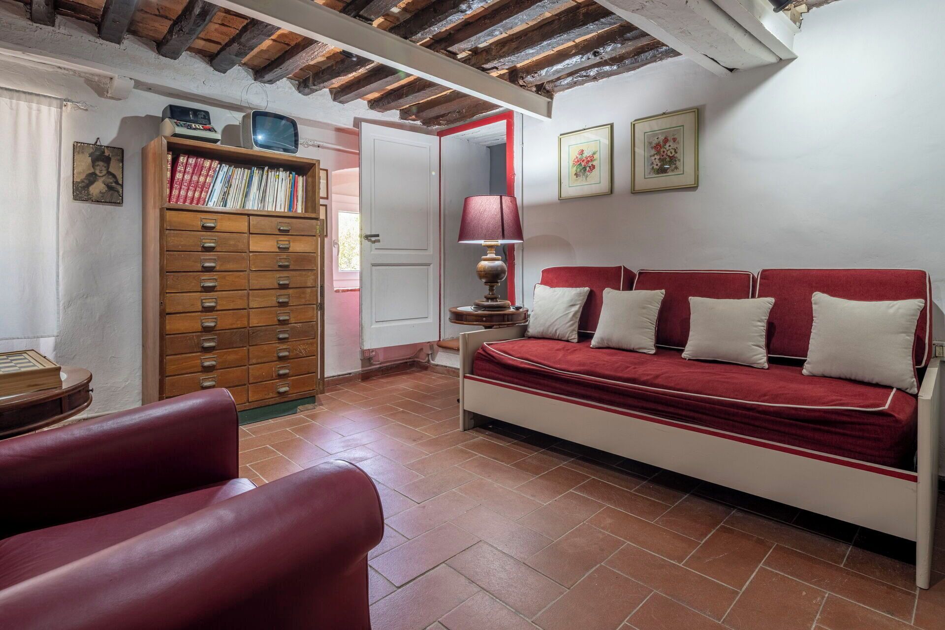 Una habitación con un sofá rojo, un armario de madera, una lámpara roja y suelo de baldosas.