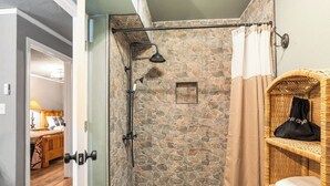 Une cabine de douche en pierre avec un pommeau de douche à main, un petit tableau encadré et une étagère en osier avec un sac.