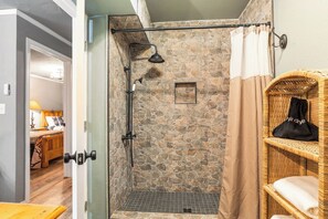 A stone shower enclosure with a handheld showerhead, a small framed picture, and a wicker shelf with a bag. - Salt Block - The Rustic 1 | Cozy 2BR in Bandera (Bandera)