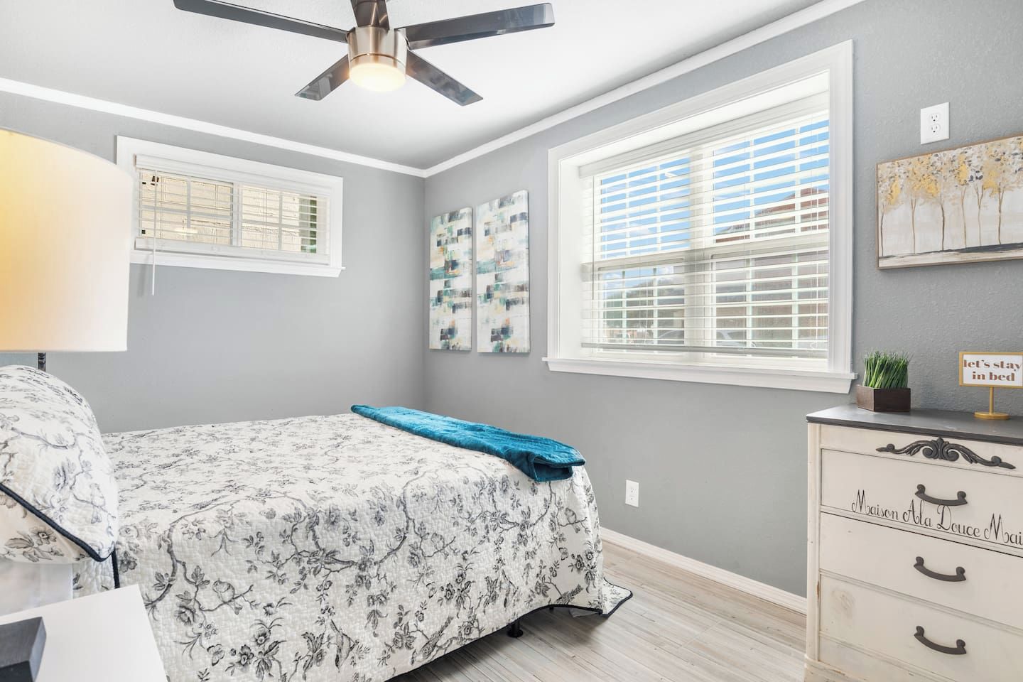 A bedroom with a bed, a dresser, a ceiling fan, and two windows with blinds.