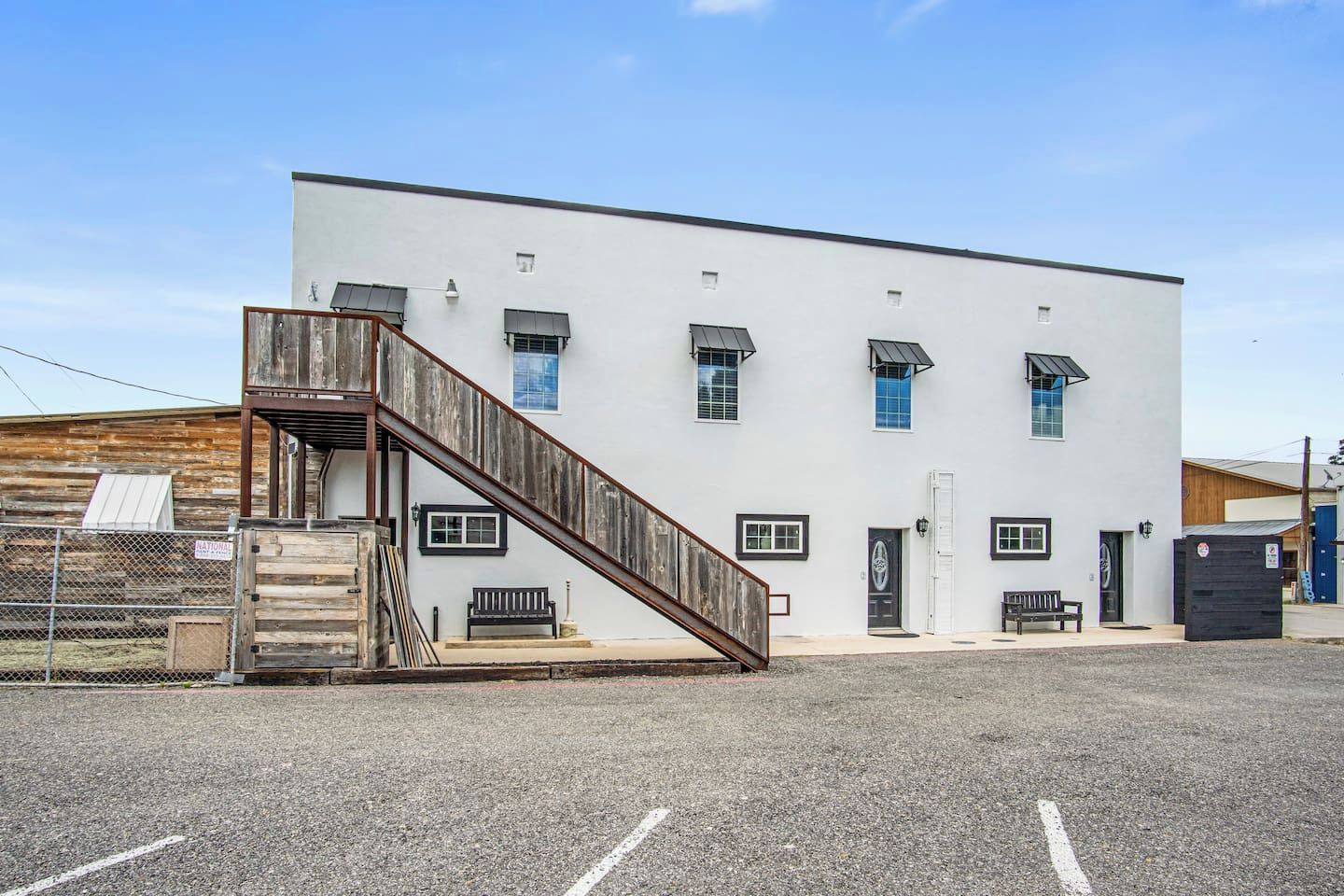 A building with a wooden staircase leading to a door, a fenced area, and a parking lot.