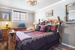 Ein Schlafzimmer mit einem rustikalen hölzernen Kopfteil, einem Bett mit gemusterten Betttextilien, einem Nachttisch mit Lampe und einem Fenster mit Jalousien.