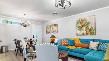 A living room with a blue sofa, a dining table with chairs, a chandelier, and artwork on the walls.