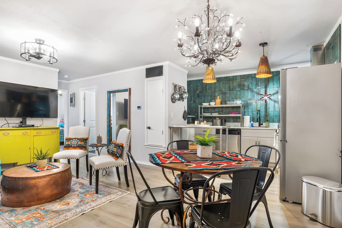 A modern kitchen with a central island, a dining area with a patterned tablecloth, and a living area with a television and colorful decor.