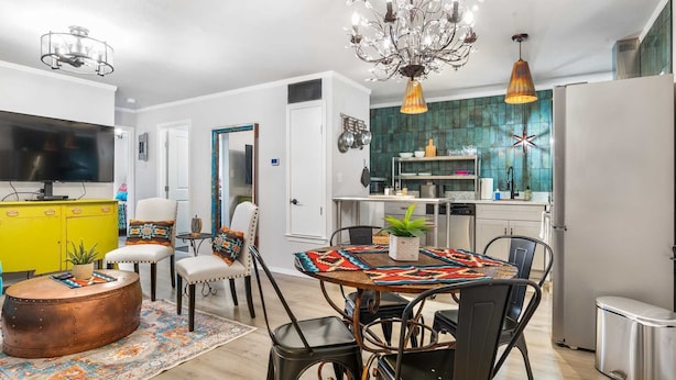 Een moderne keuken met een blauwgroene achterwand, een eethoek met een tafelkleed met patroon, en een woonkamer met een televisie en een geel kastje.