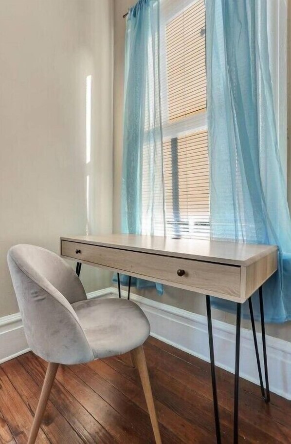 A corner desk with a light wooden finish, a grey upholstered chair, and a window with blue sheer curtains.