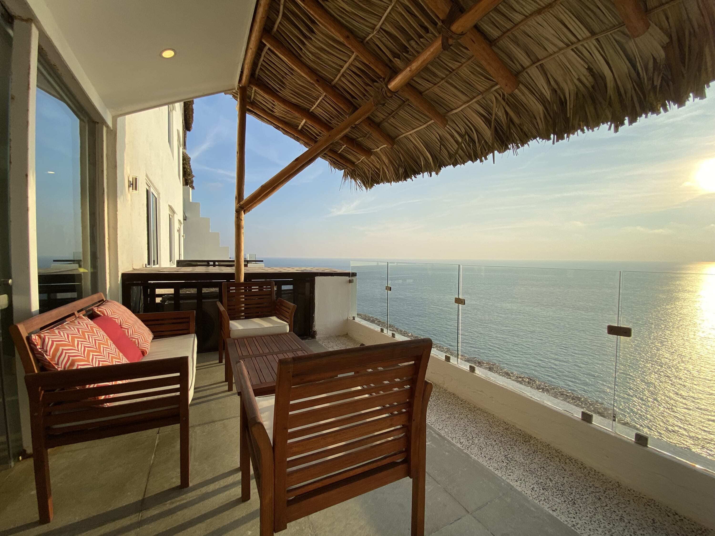 A balcony with wooden furniture, a thatched roof, and a view of the sea.