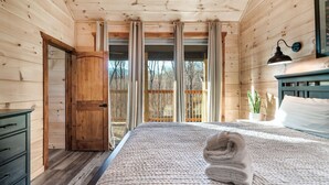 Un dormitorio de cabaña de madera con una cama, un armario de madera, una ventana con cortinas y una mesita de noche con lámpara.
