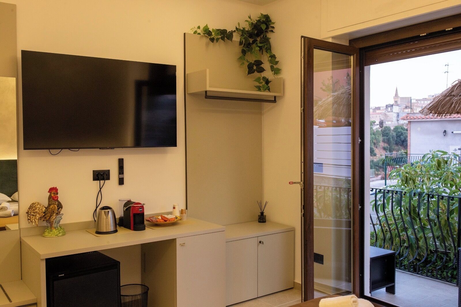 A modern hotel room with a flat-screen TV, a coffee machine, a small refrigerator, a sink, and a balcony with a view of the city.