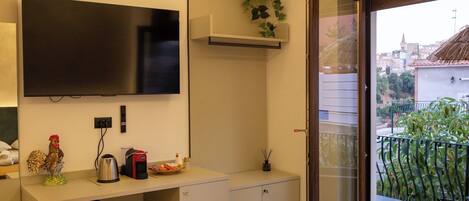 A modern hotel room with a flat-screen TV, a coffee machine, a small refrigerator, a sink, and a balcony with a view of the city.
