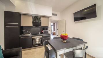 A modern kitchen with a dining area, a flat-screen TV, and a bowl of fruit on the table.