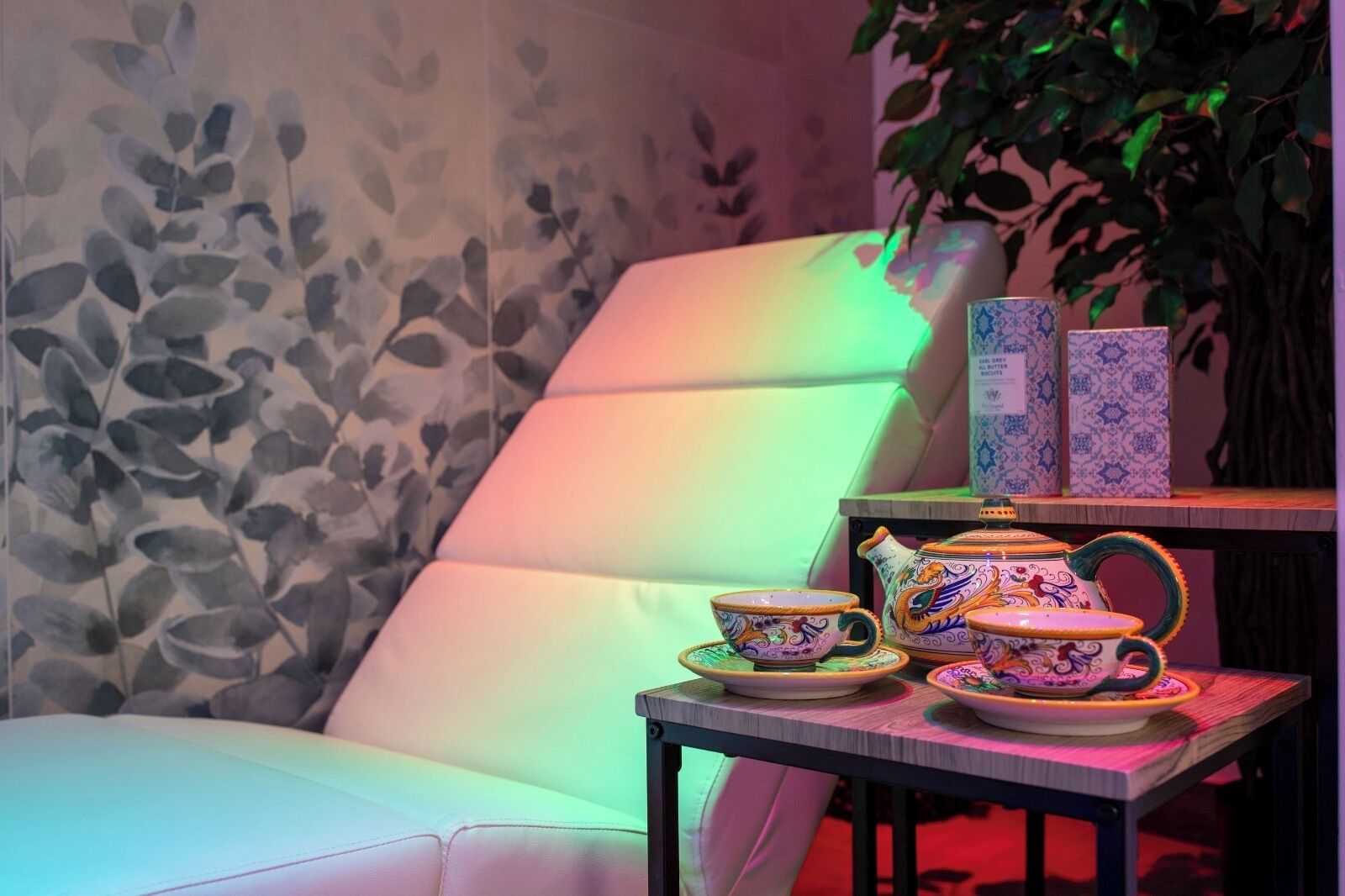 Une pièce dont les murs sont recouverts d’un papier peint à motifs floraux, avec un fauteuil blanc rembourré, une table sur laquelle se trouvent une théière, des tasses et une boîte.