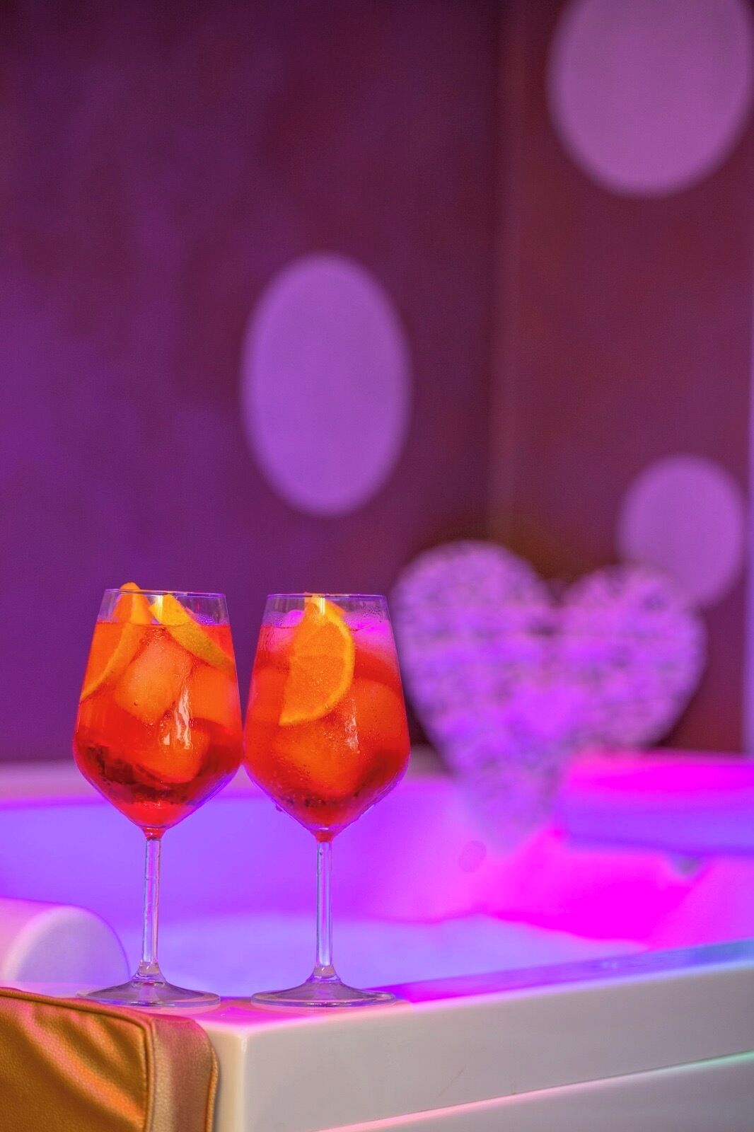 Dois copos de uma bebida vermelha com fatias de laranja, sobre uma superfície com decoração em formato de coração ao fundo.