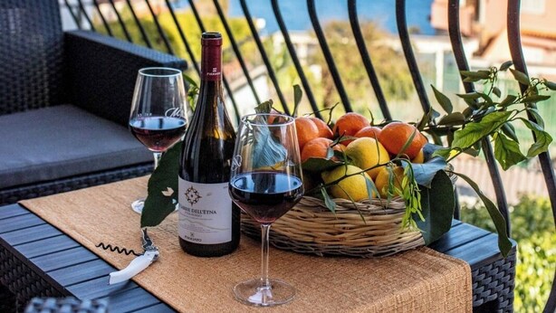 Un balcon avec une table en osier, mise pour deux, agrémentée d’une bouteille de vin, de deux verres et d’un panier de fruits.