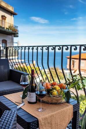 A balcony with a wicker table set for two, featuring a bottle of wine, two glasses, and a basket of fruit. - SUITE APARTMENT TAORMINA (Taormina)