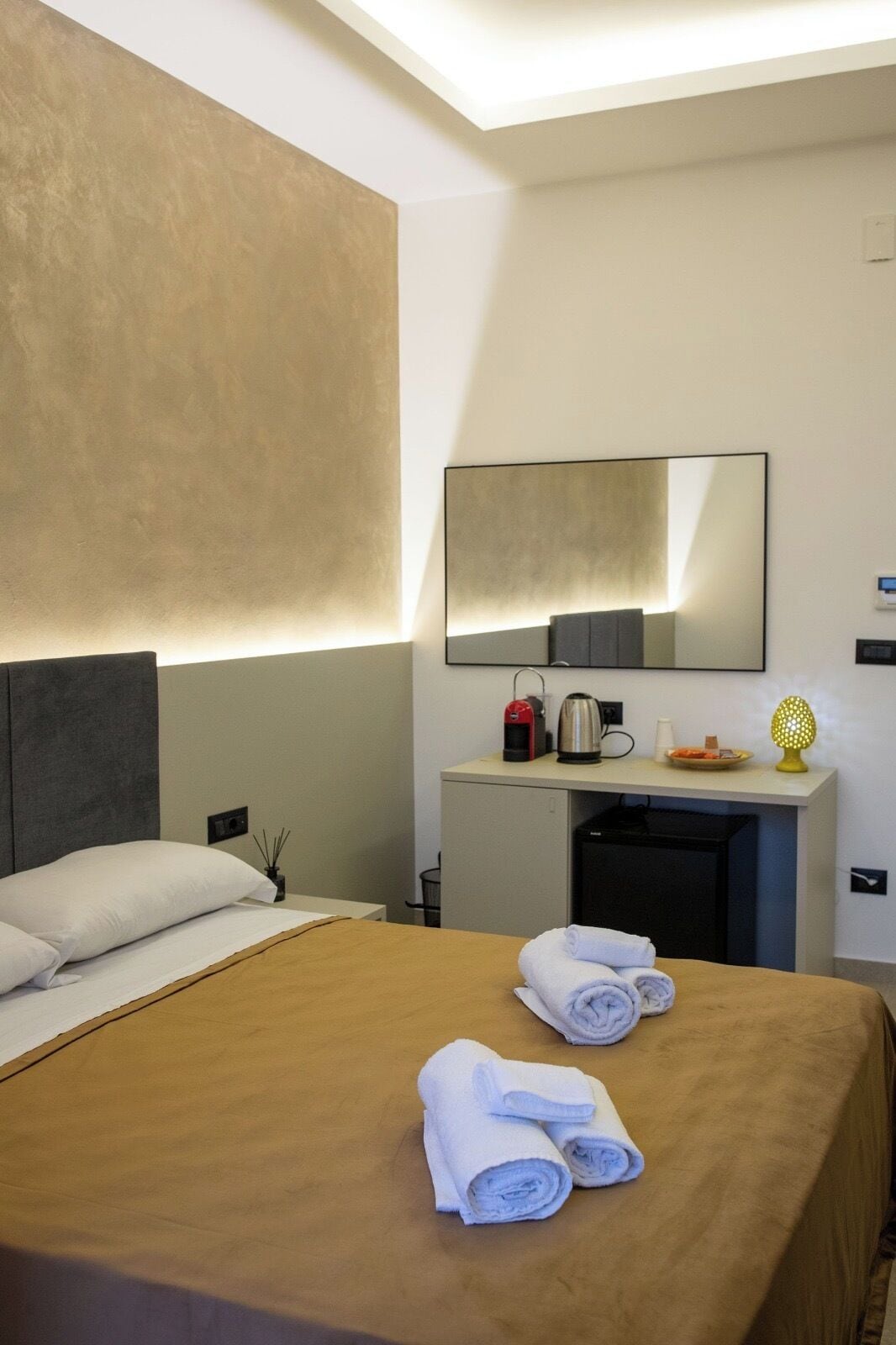 A hotel room with a bed, a desk with a kettle, a lamp, and a mirror.