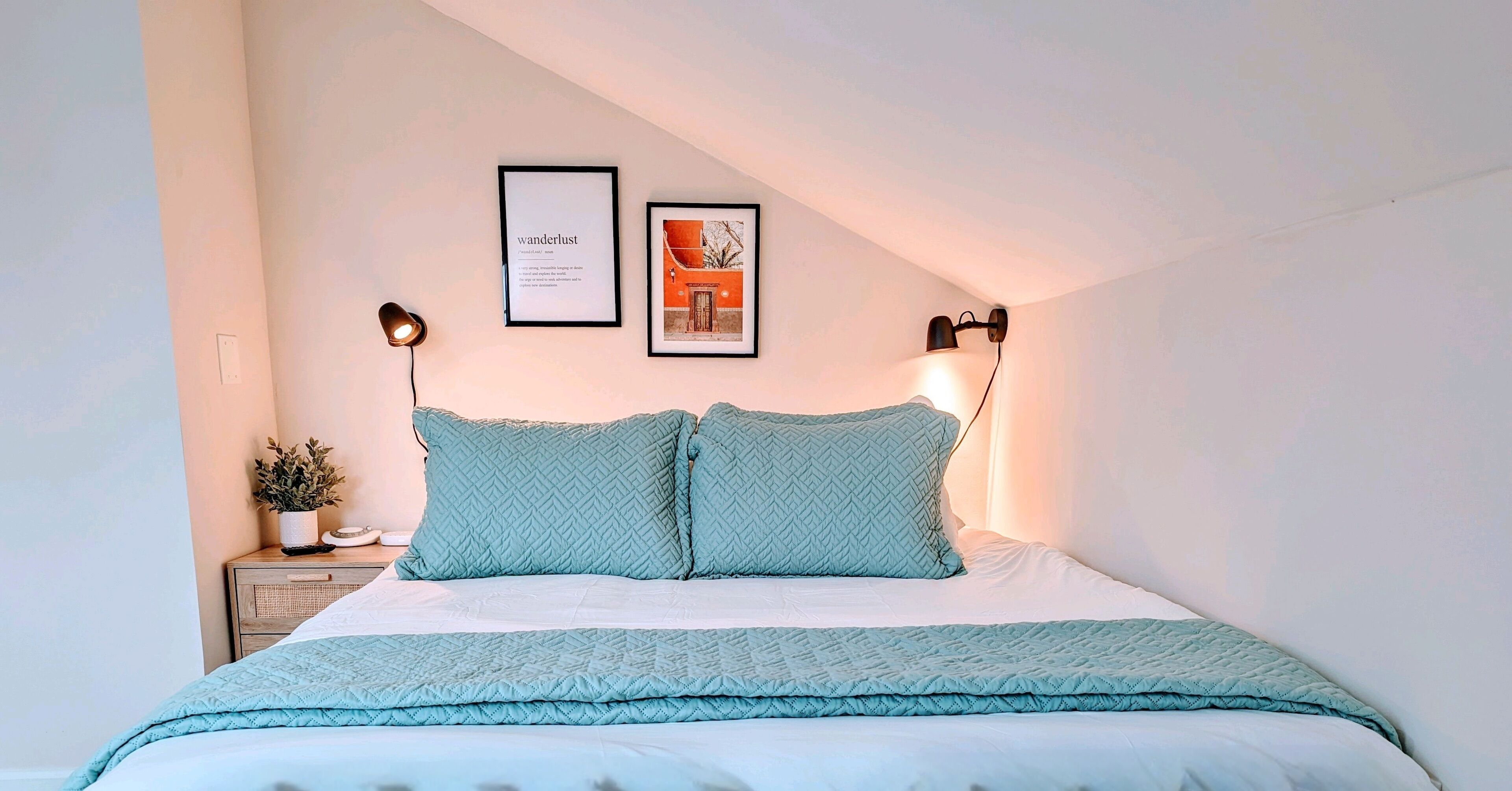 A neatly made bed with a light blue quilt and pillows, a small bedside table with a plant, and two framed pictures on the wall.