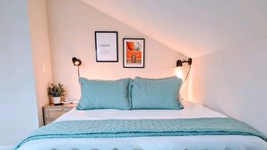 A neatly made bed with a light blue quilt and pillows, a small bedside table with a plant, and two framed pictures on the wall. - Unique Little Charmer in Downtown Frederick

 (Frederick)