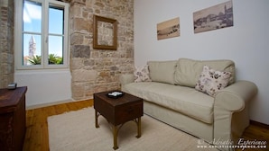 Une pièce avec un mur en pierre, une fenêtre donnant sur une tour, un canapé beige et deux tableaux encadrés accrochés au mur.