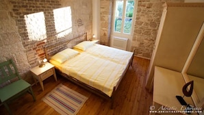 Une chambre avec un mur en briques, un lit recouvert d’une courtepointe à rayures, une table de chevet avec une lampe, une chaise et une porte donnant sur un jardin.