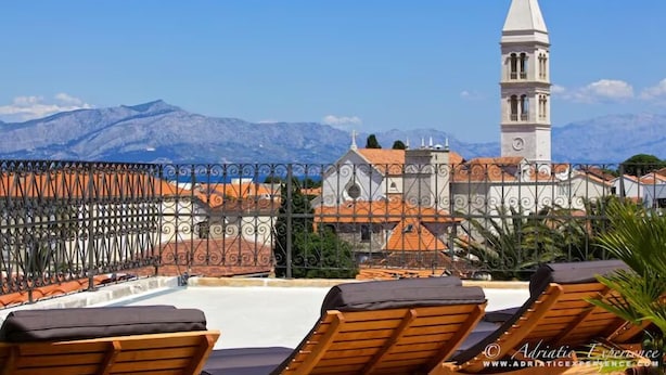 Une terrasse sur le toit, dotée de chaises longues, d’une clôture décorative et offrant une vue sur un bâtiment historique, avec un clocher et des montagnes en arrière-plan.