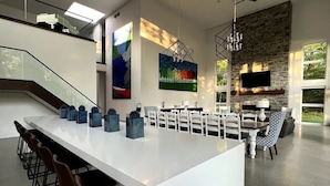 A modern kitchen with a large island, a dining area with chairs, and a stone fireplace. - Muskoka Modern Luxury Lake House Cottage (Port Carling)