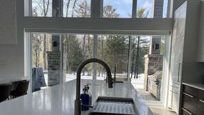 A modern kitchen with a large island, a stainless steel sink, and a view of a snowy landscape through large windows. - Muskoka Modern Luxury Lake House Cottage (Port Carling)