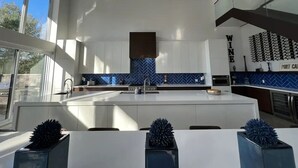 A modern kitchen with white cabinets, a blue backsplash, and a central island. There are three skylights and a sign that reads "WINE". - Muskoka Modern Luxury Lake House Cottage (Port Carling)