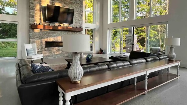 A spacious living room with a stone fireplace, a large flat-screen TV, a long wooden bench, and large windows offering a view of the outdoors. - Muskoka Modern Luxury Lake House Cottage (Port Carling)