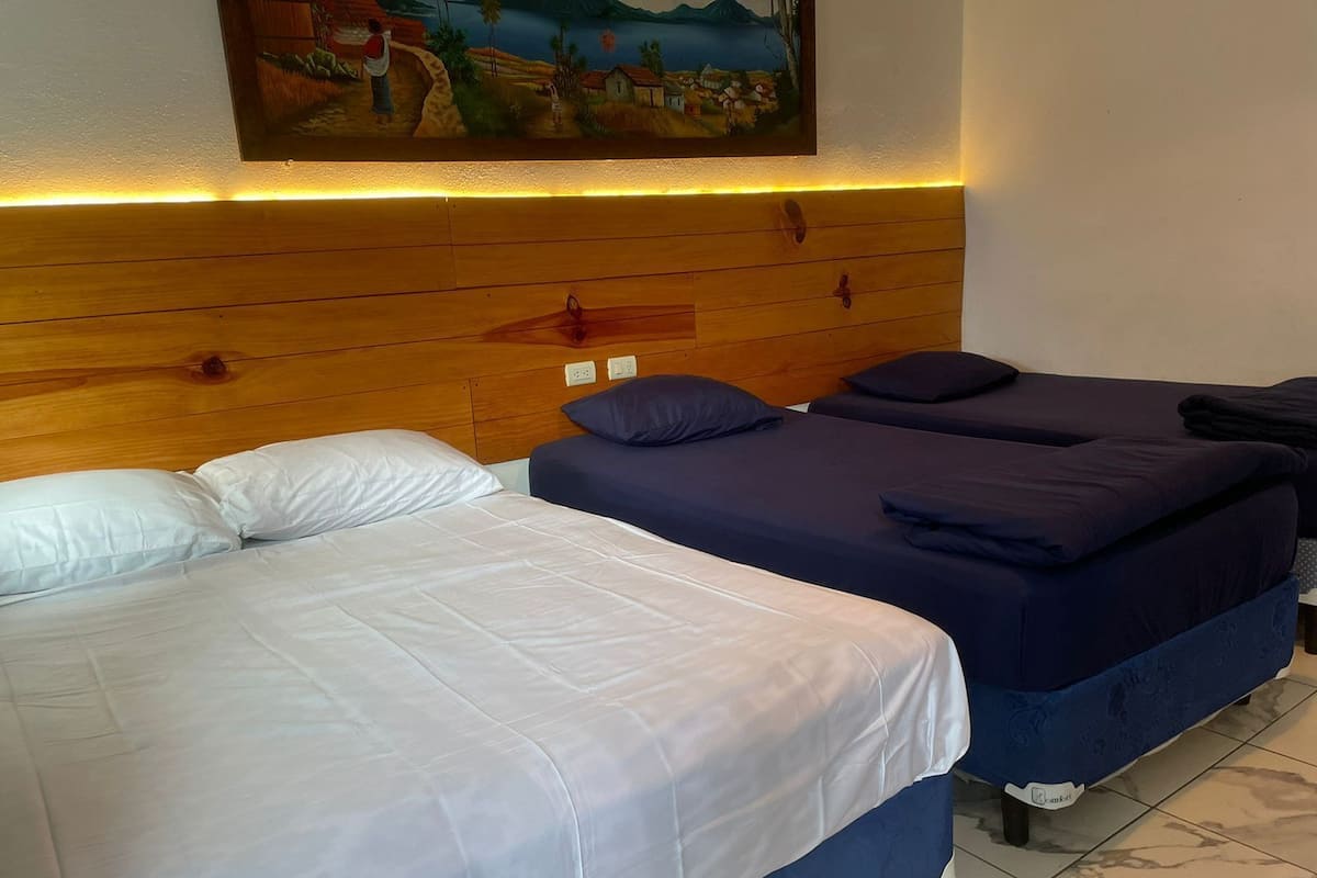 Habitación de hotel con dos camas, una con ropa blanca y la otra con ropa de color azul oscuro. Tiene un cabecero de madera y un cuadro encima.