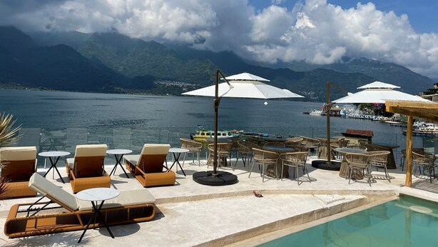 Zona junto a la piscina con sillones de descanso y sombrillas, con vistas a un cuerpo de agua tranquilo y montañas.