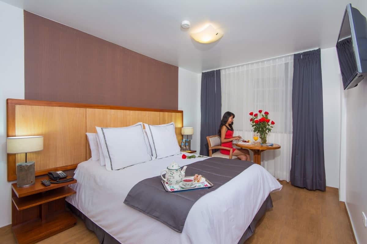 A hotel room with a large bed, bedside tables, a TV, and a dining area with a table and chairs.