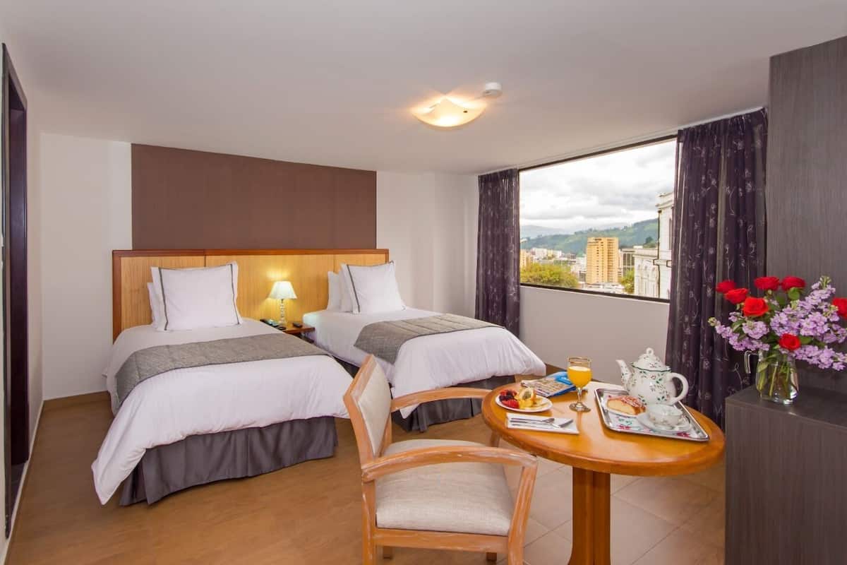 A hotel room with two beds, a dining table with breakfast items, and a city view through the window.