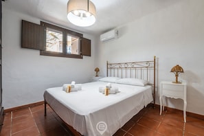 A bedroom with a bed, a nightstand, a lamp, a window with shutters, and an air conditioning unit.
