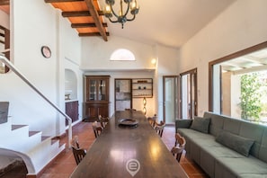 A spacious dining area with a long wooden table, surrounded by chairs, and a sofa. A staircase with a wooden railing is visible on the left.