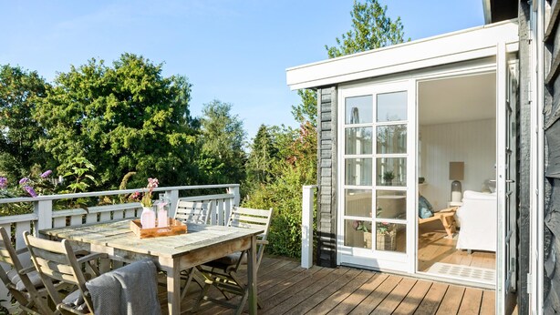 A wooden deck with a table and chairs, a small white building with a sliding glass door, and a garden with trees and flowers. - Amazing home in Hornbæk with WiFi (Hornbæk)