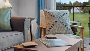 A modern living room with a grey sofa, a wooden coffee table with a book, a patterned throw pillow, and a view of a green landscape through a large window.