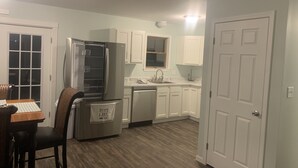A compact kitchen with white cabinets, a stainless steel refrigerator, a dishwasher, and a dining area with a table and chairs. - Cozy Lake House near Pymatuning Lake (Jamestown)