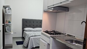 Une cuisine compacte équipée d’une cuisinière à gaz, d’un four encastré et d’un évier. On y trouve également un petit réfrigérateur et un four à micro-ondes posés sur le plan de travail. On aperçoit un lit avec une tête de lit grise au fond de la pièce.