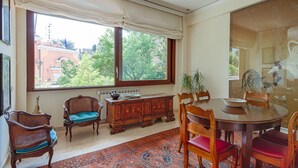 A dining area with a round table, chairs, a wooden cabinet, and a large window with a view of trees and buildings. - Parioli Luxury Terrace (Roma)