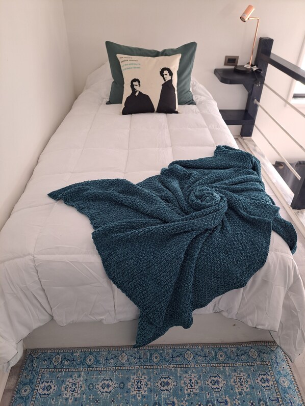 Una cama con edredón blanco, una manta turquesa y un cojín con el logo de una banda.